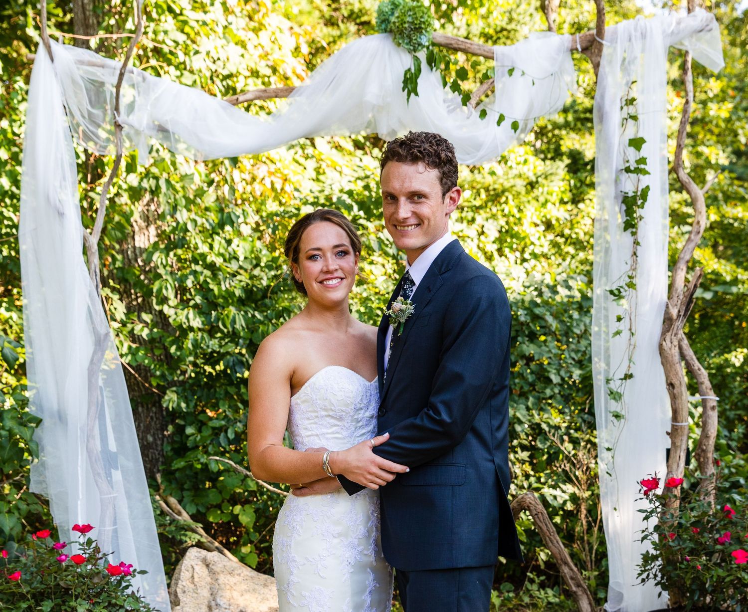 A wedding ceremony takes place under an elegant white draped canopy in a lush garden setting.