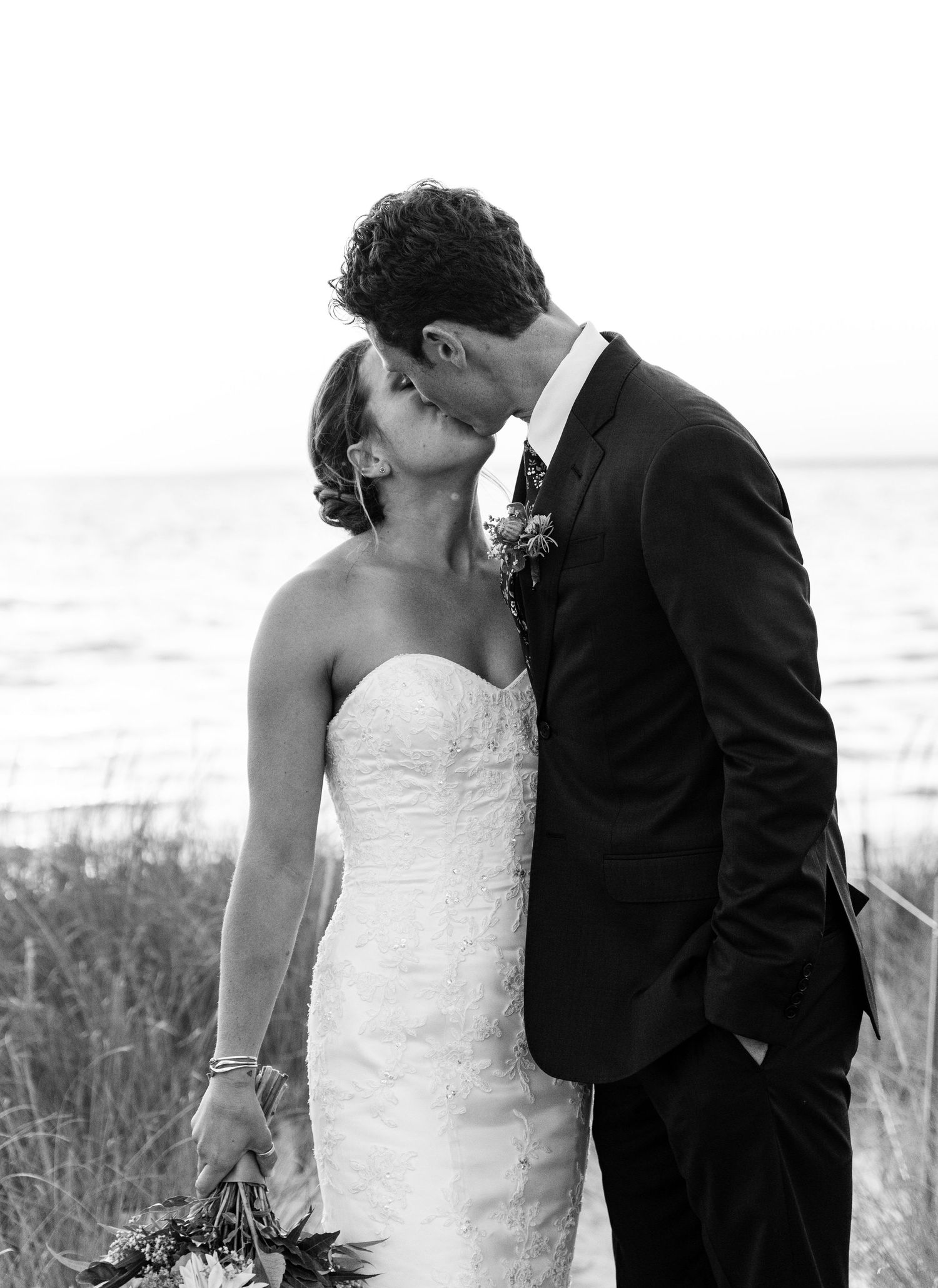 A romantic black and white wedding photograph of a couple sharing an intimate kiss by the ocean.