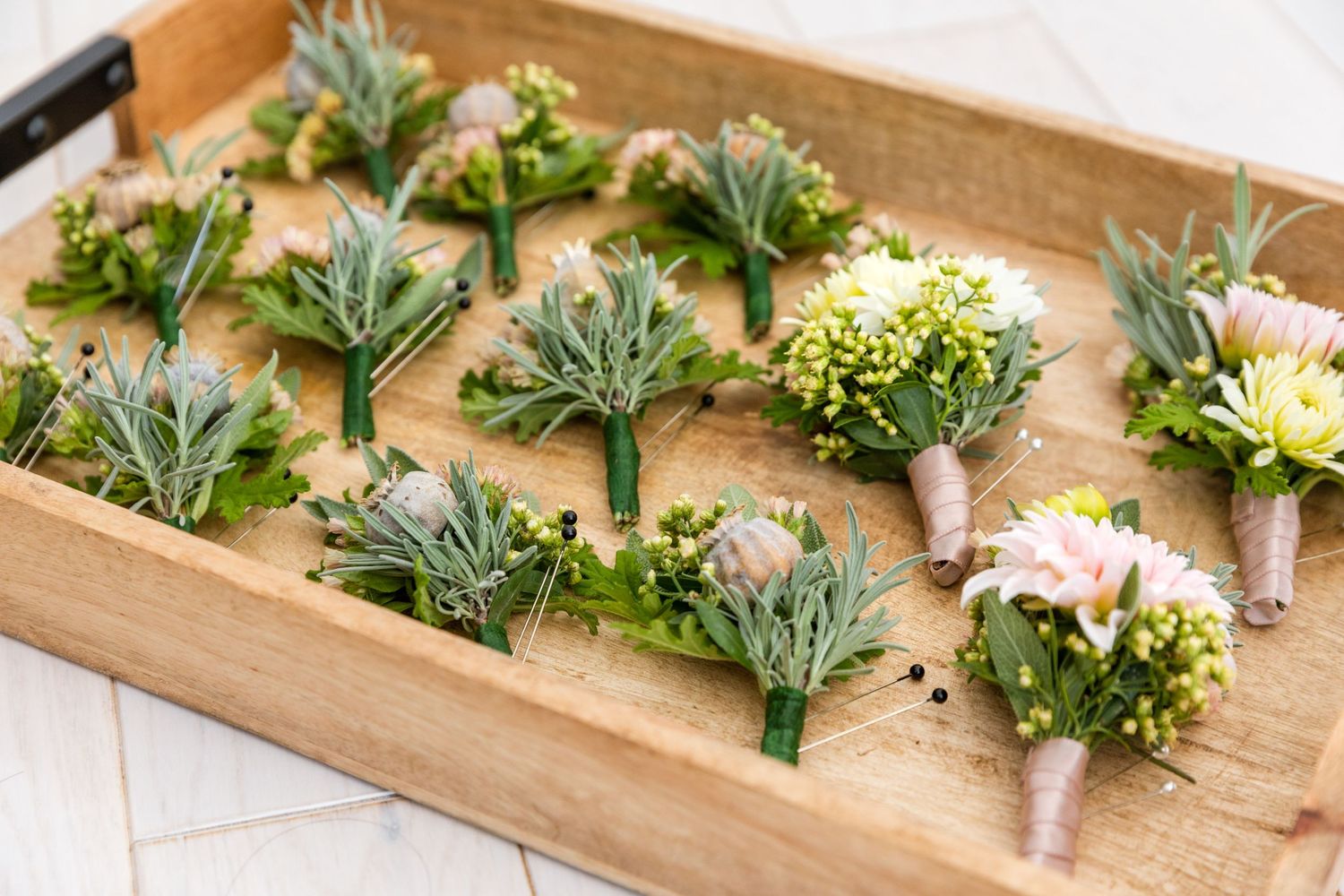 Small wedding boutonnieres with delicate white flowers and dusty miller leaves arranged on a wooden tray.