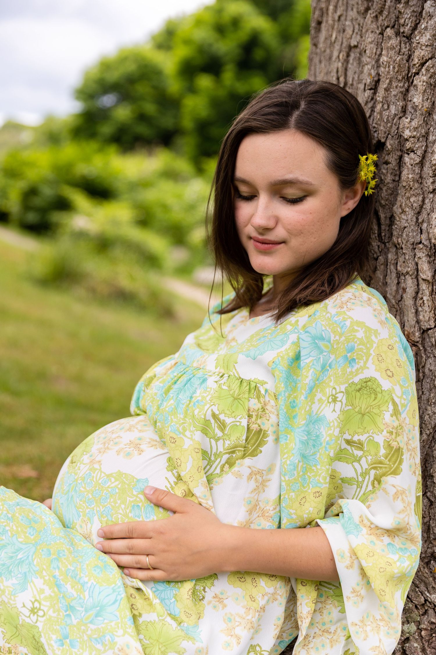 A pregnant woman in a floral yellow-green dress rests against a tree trunk outdoors with a small flower in her hair.
