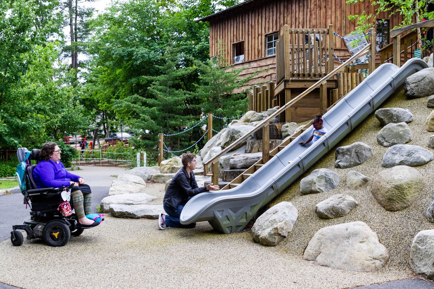 Children playing on a built-in metal slide among rocks outside a rustic wooden building at an accessible playground.