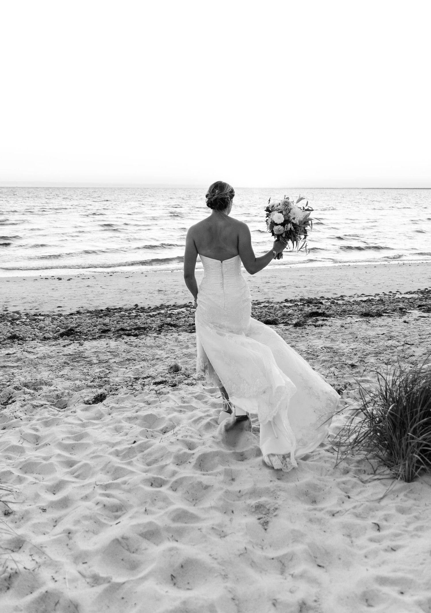 A bride in a flowing white dress stands on a sandy beach holding a bouquet while looking out at the ocean.