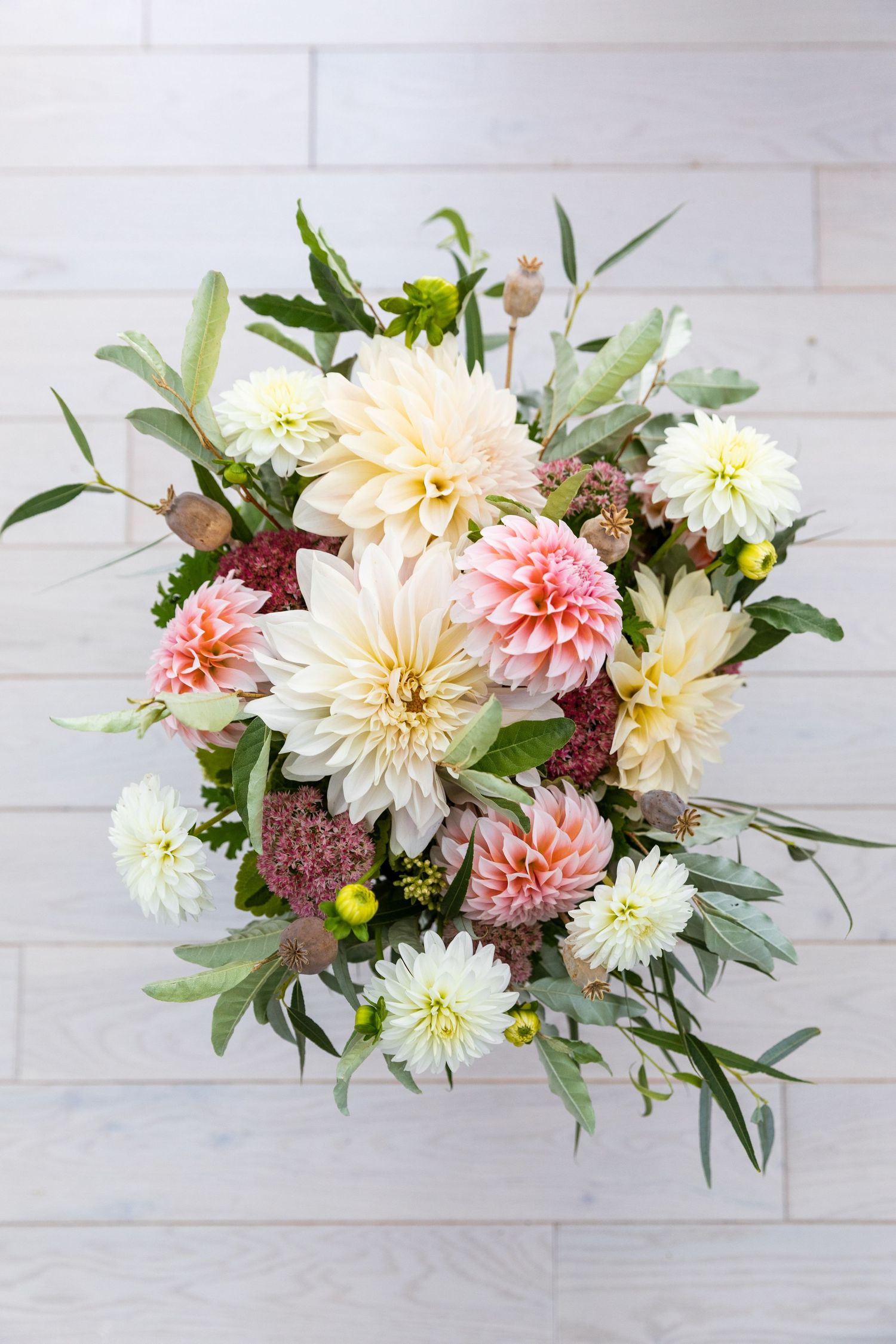 A romantic floral arrangement with peach dahlias, pink blossoms, and green foliage against a white brick wall.