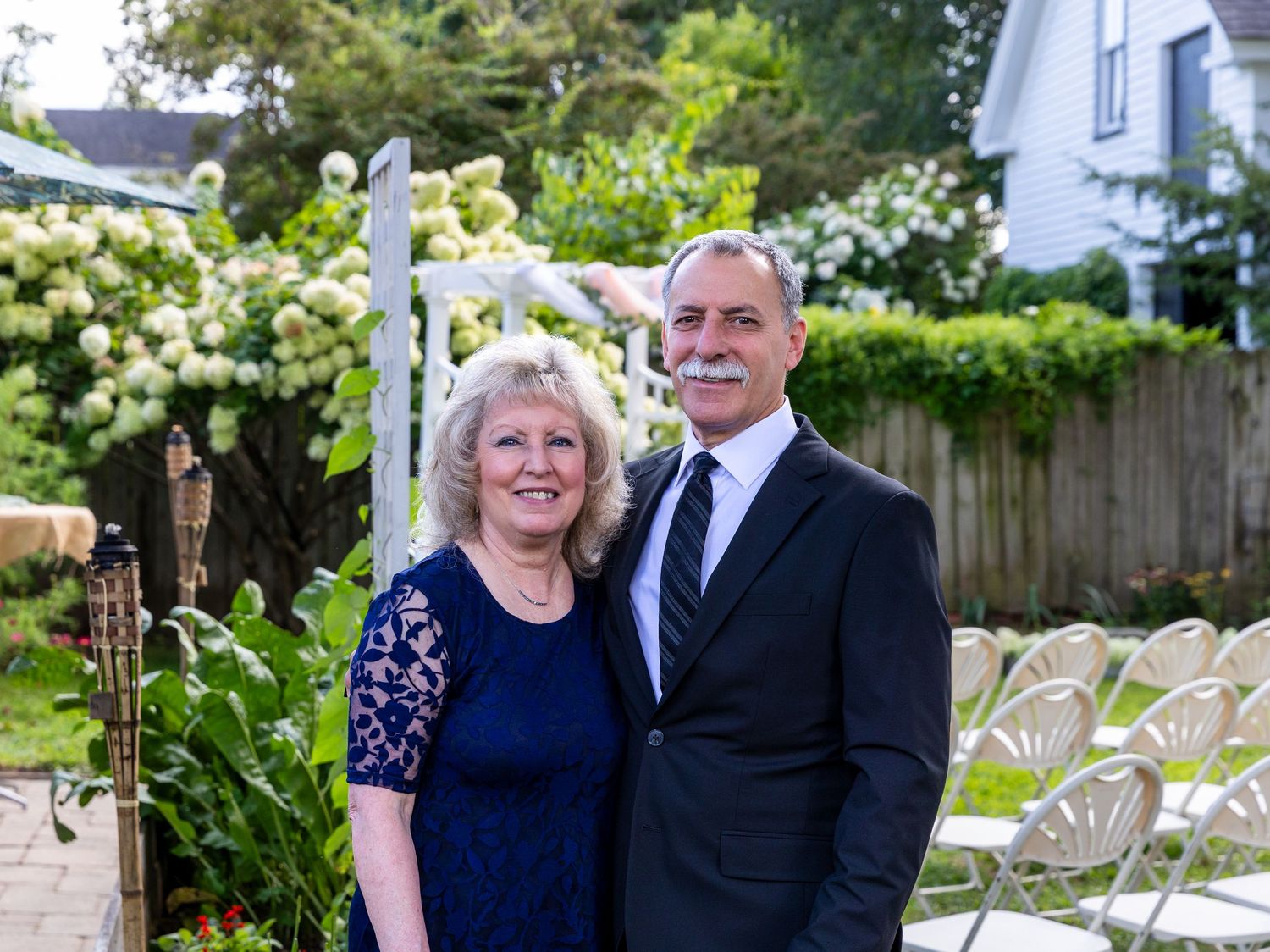 A couple dressed in formal navy and black attire poses together in an outdoor garden setting with flowers and greenery.