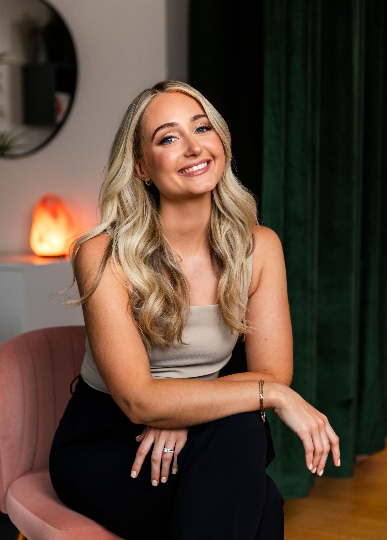 A woman in a gray tank top and black pants sits on a pink chair, smiling warmly in a room with green curtains and mood lighting.