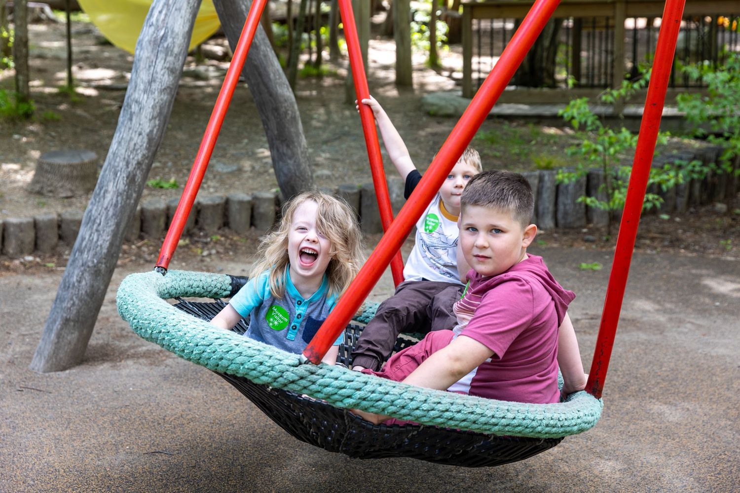 Two kids smiling and laughing while riding together on a green basket swing at an outdoor playground.
