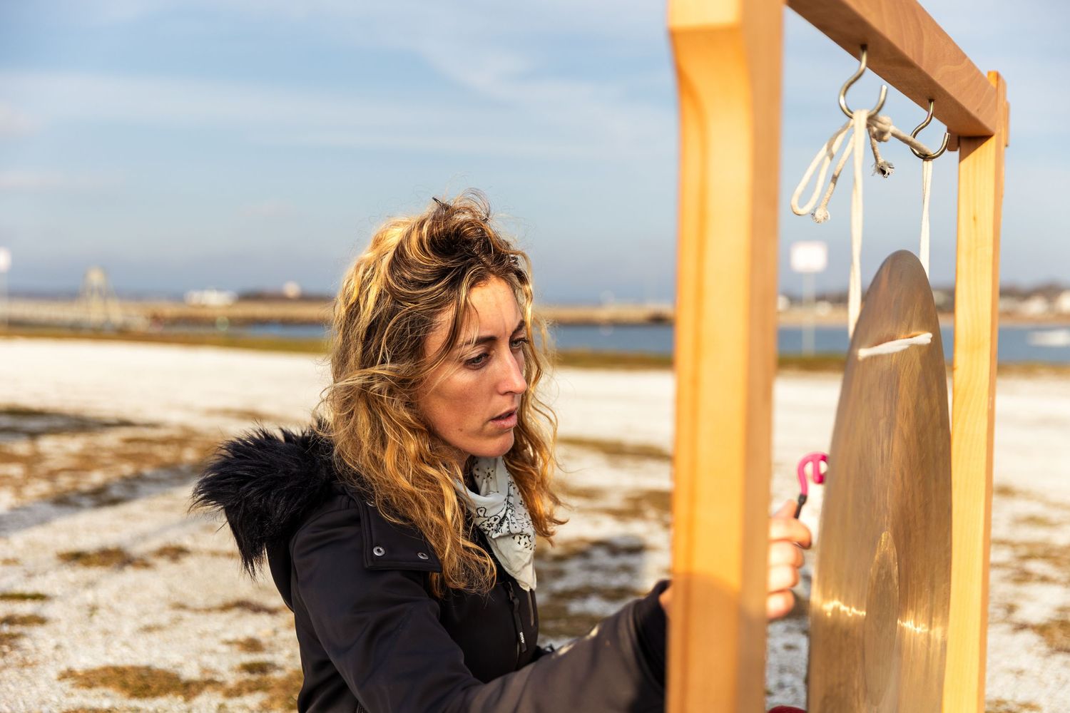 Person in a black winter coat adjusts instruments on a wooden outdoor frame in a beachy coastal setting.