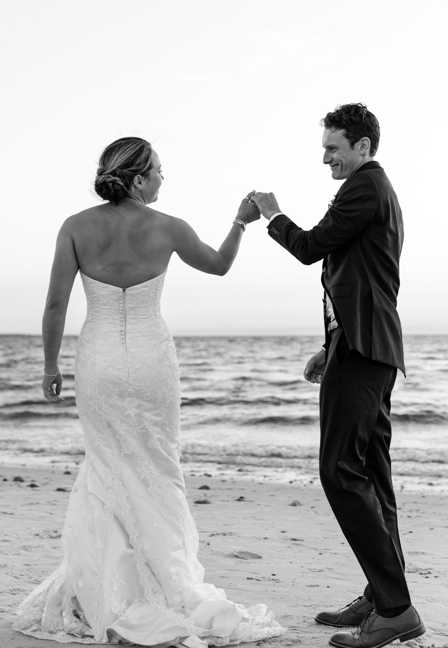 A couple shares a celebratory fist bump on the beach, with the bride in white gown and groom in dark suit.