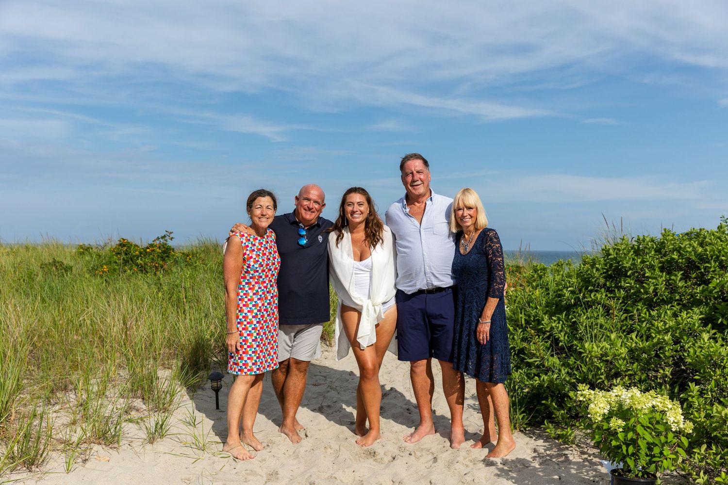 Panoramic view of family group walking on sand dunes at a coastal beach with blue sky and green vegetation.