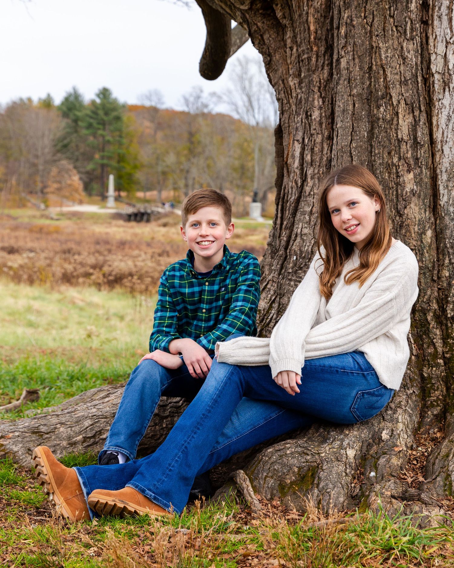 Two people sit casually against a large tree trunk in a peaceful autumn field setting.
