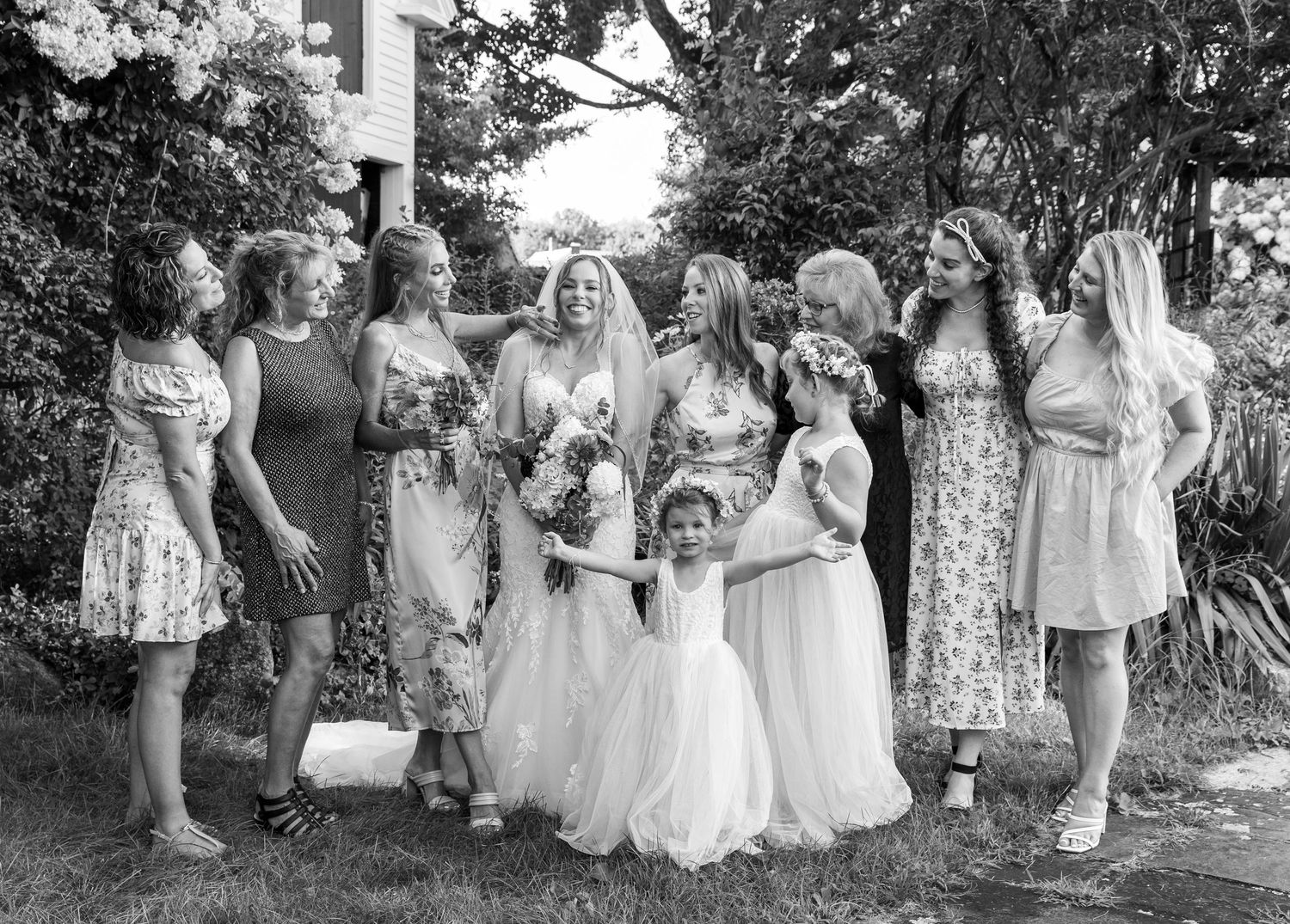 Black and white photo of a wedding party in a garden setting with bridesmaids in floral dresses gathered together.