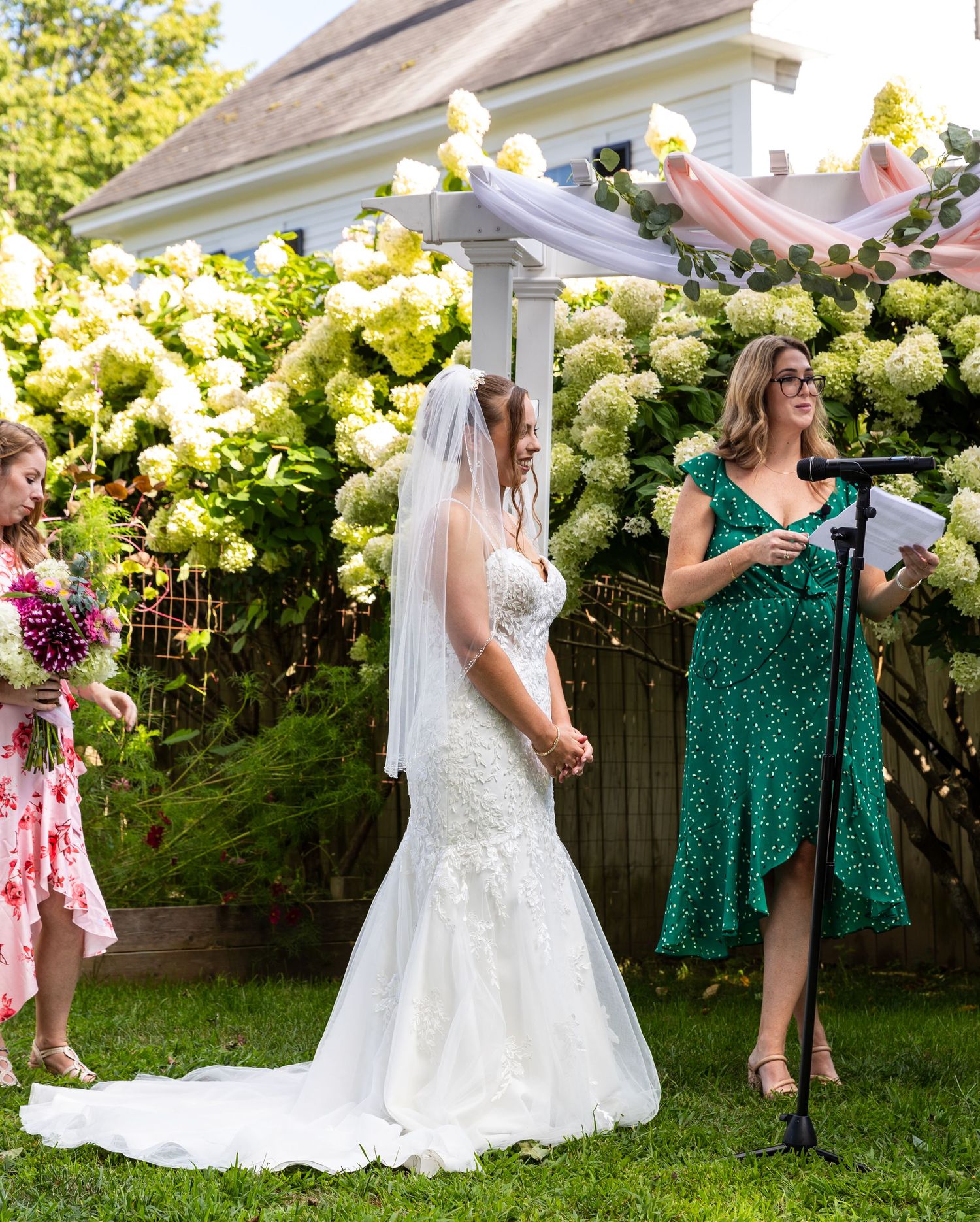 Wedding party in green dresses and white gown toasting with drinks in a backyard garden setting on a sunny day.