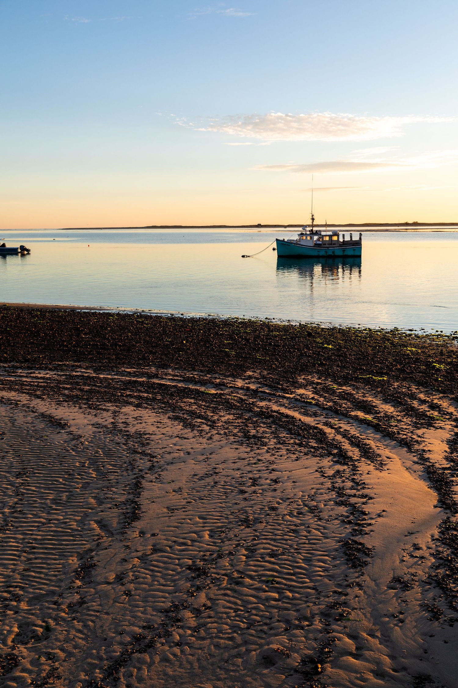 Peaceful sunset harbor scene with fishing boat silhouetted against golden sky and rippled sand beach.
