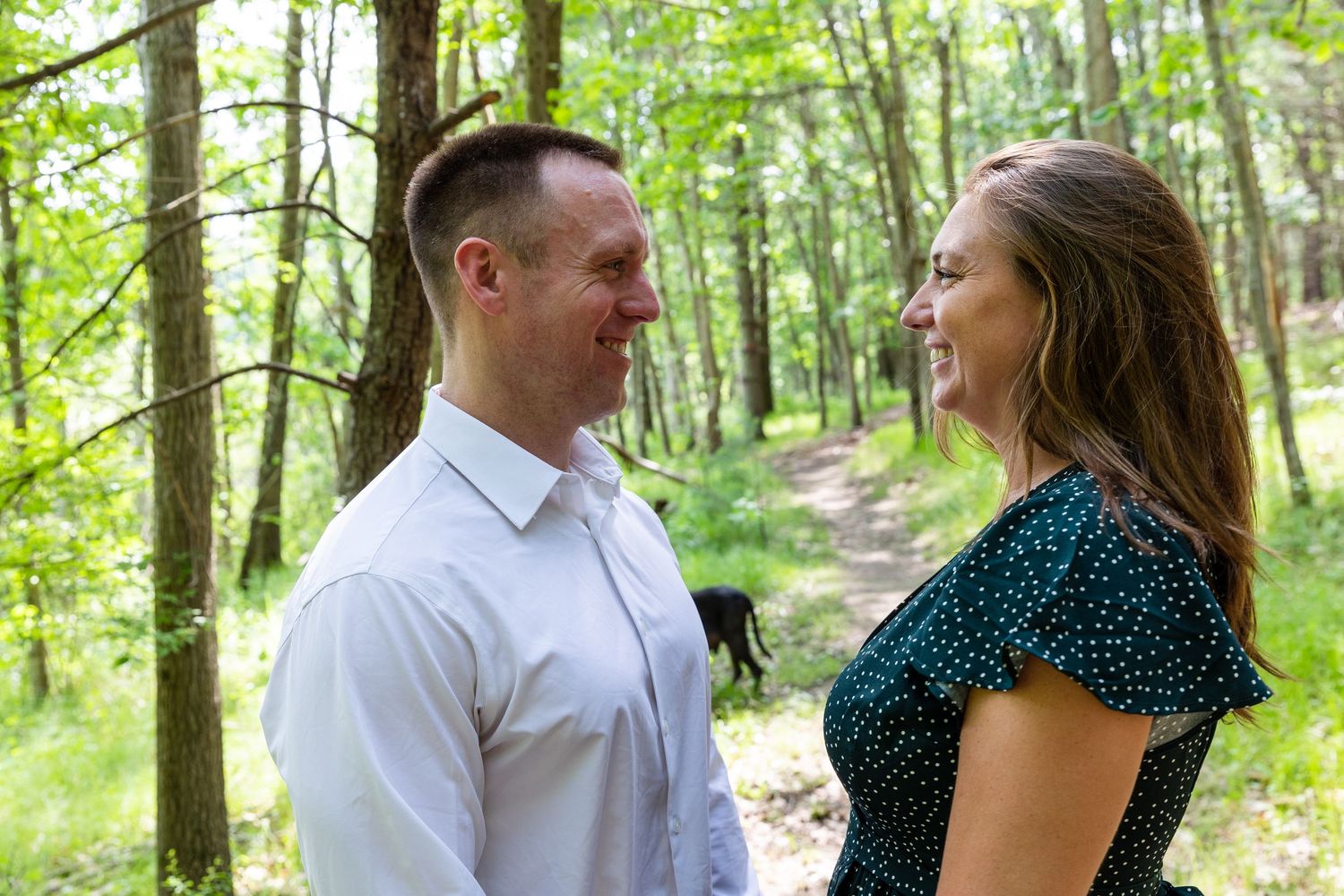 A couple shares a joyful moment together on a sunlit forest trail surrounded by green trees.