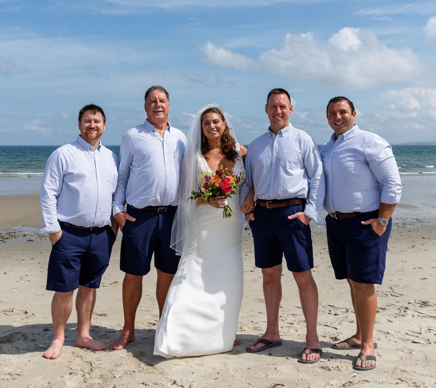 Wedding party members line up along a sunny beach in crisp white and navy attire for fun group photo poses.