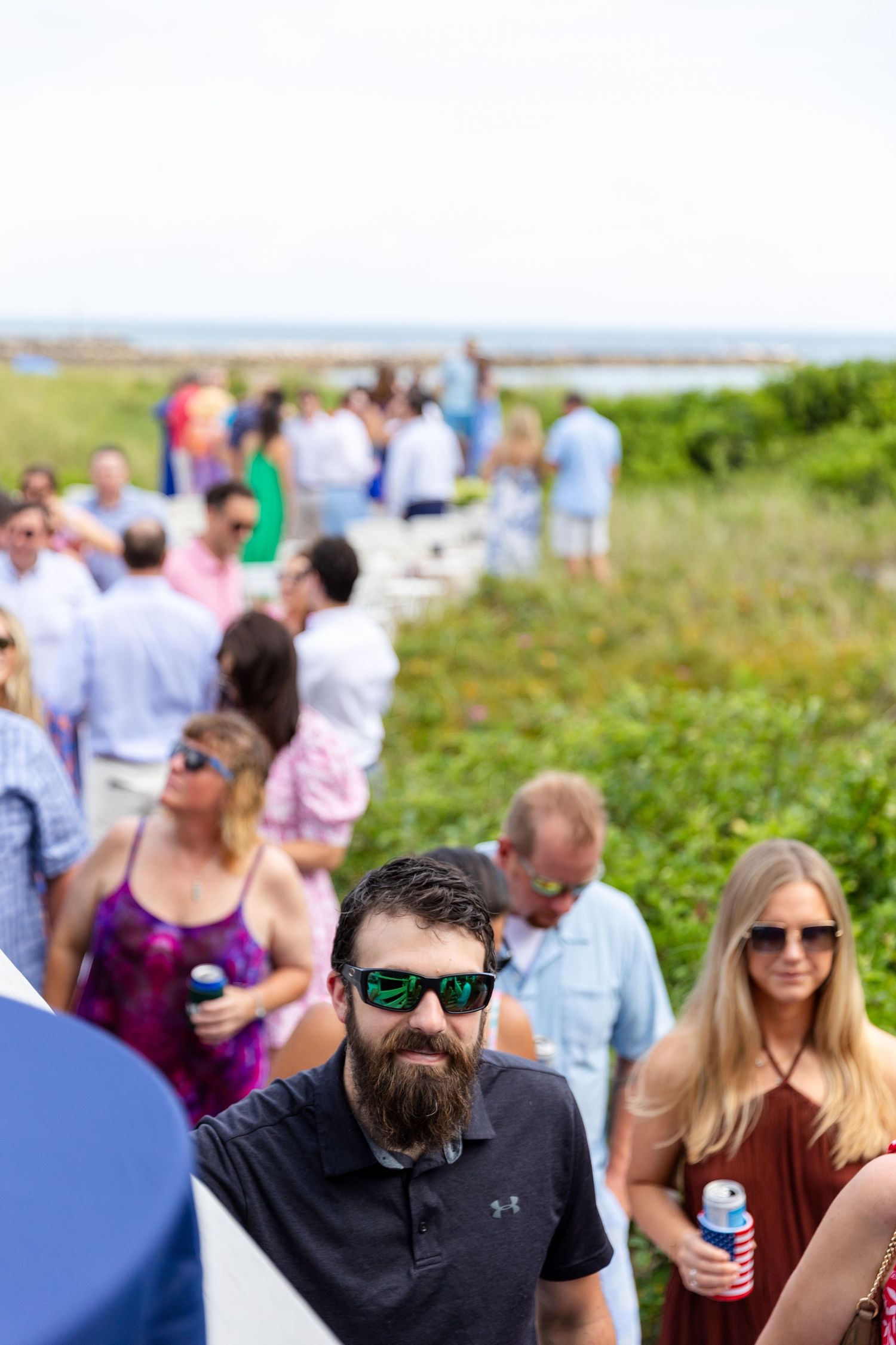 Group of people gathering outdoors on a grassy hill near the coast, holding drinks and wearing sunglasses.