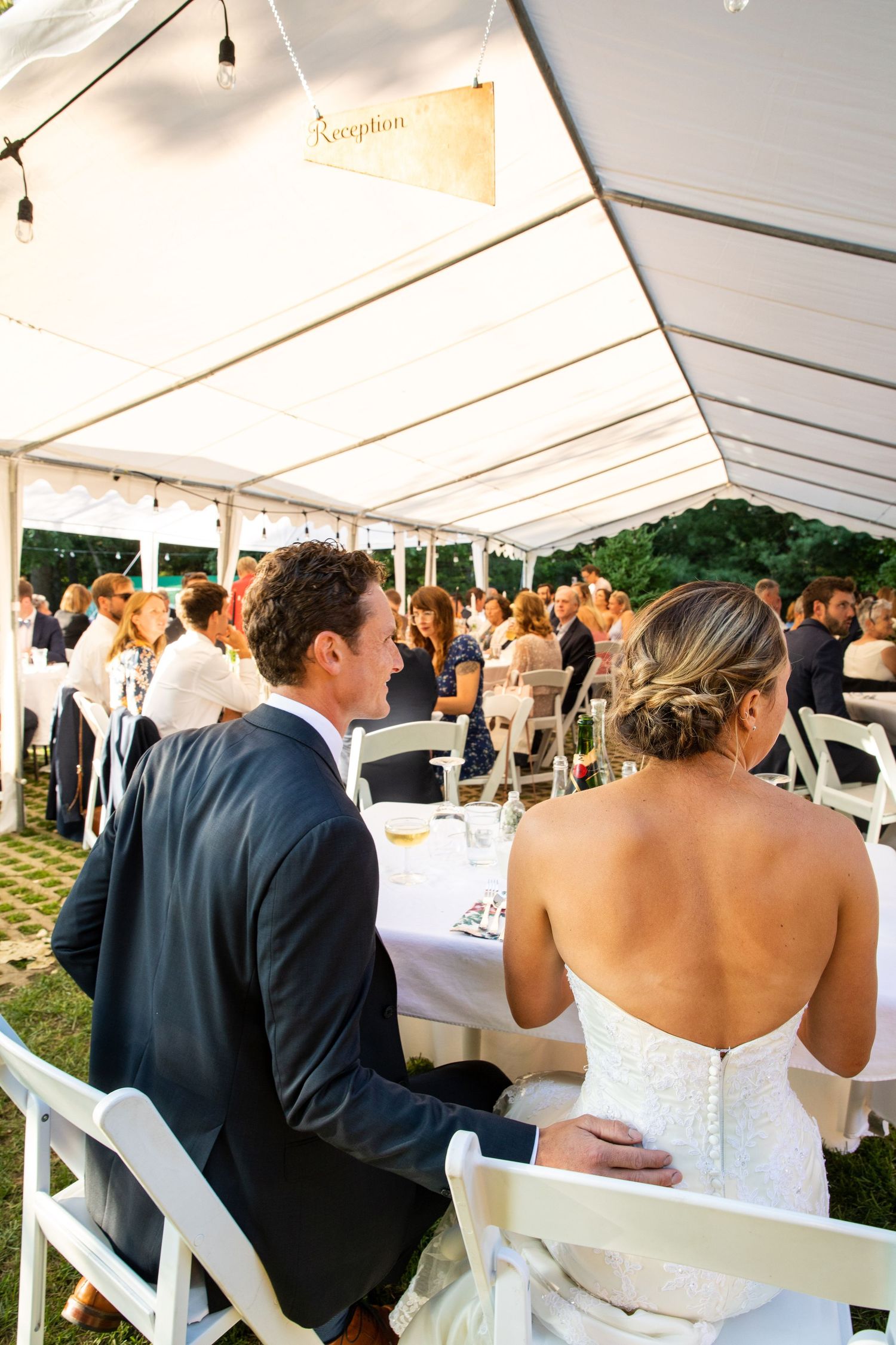 Outdoor wedding reception under white tent with elegantly set tables and hanging string lights during sunset.