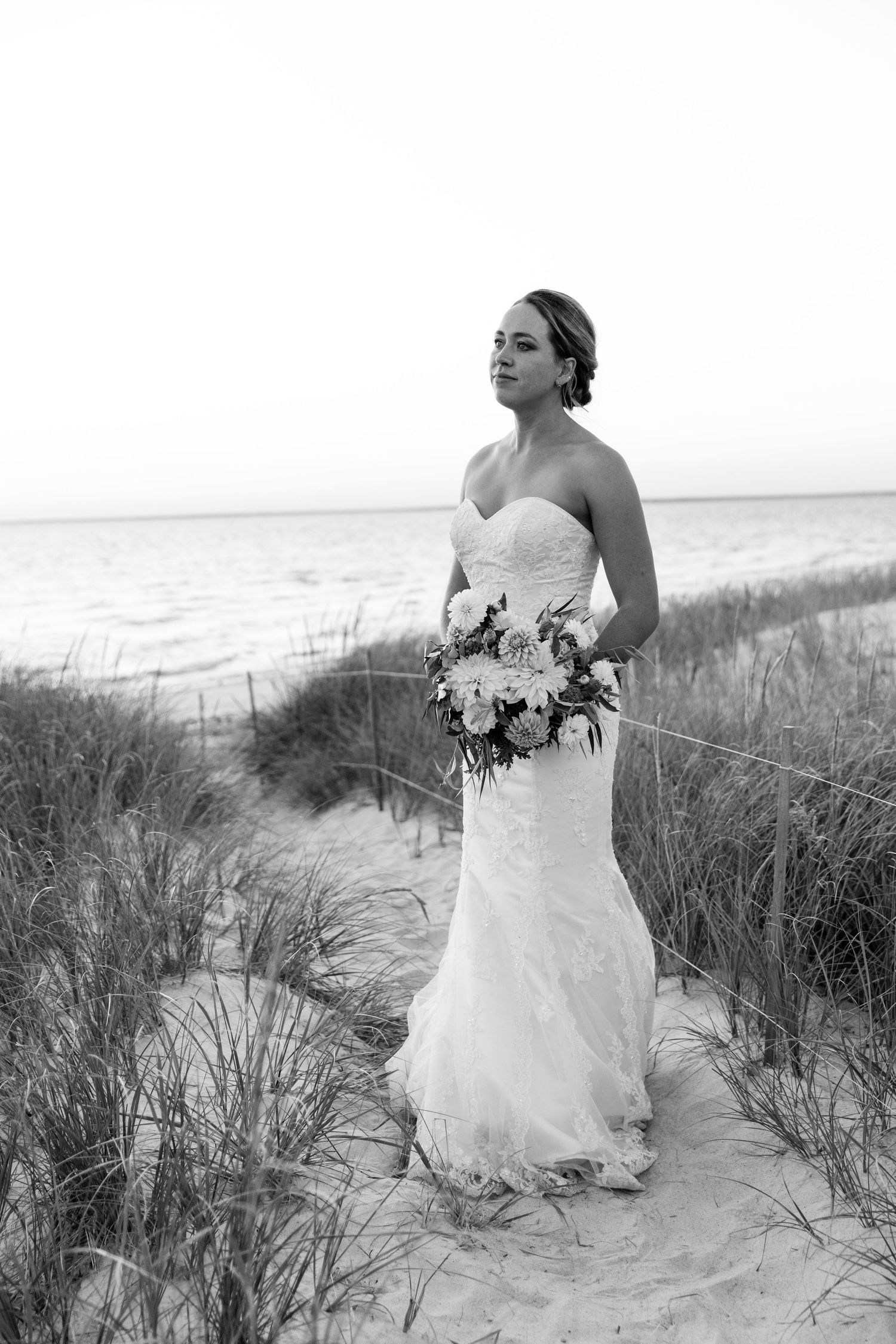 A bride in a flowing white wedding dress poses on a sandy beach dune among tall grass at the seaside.
