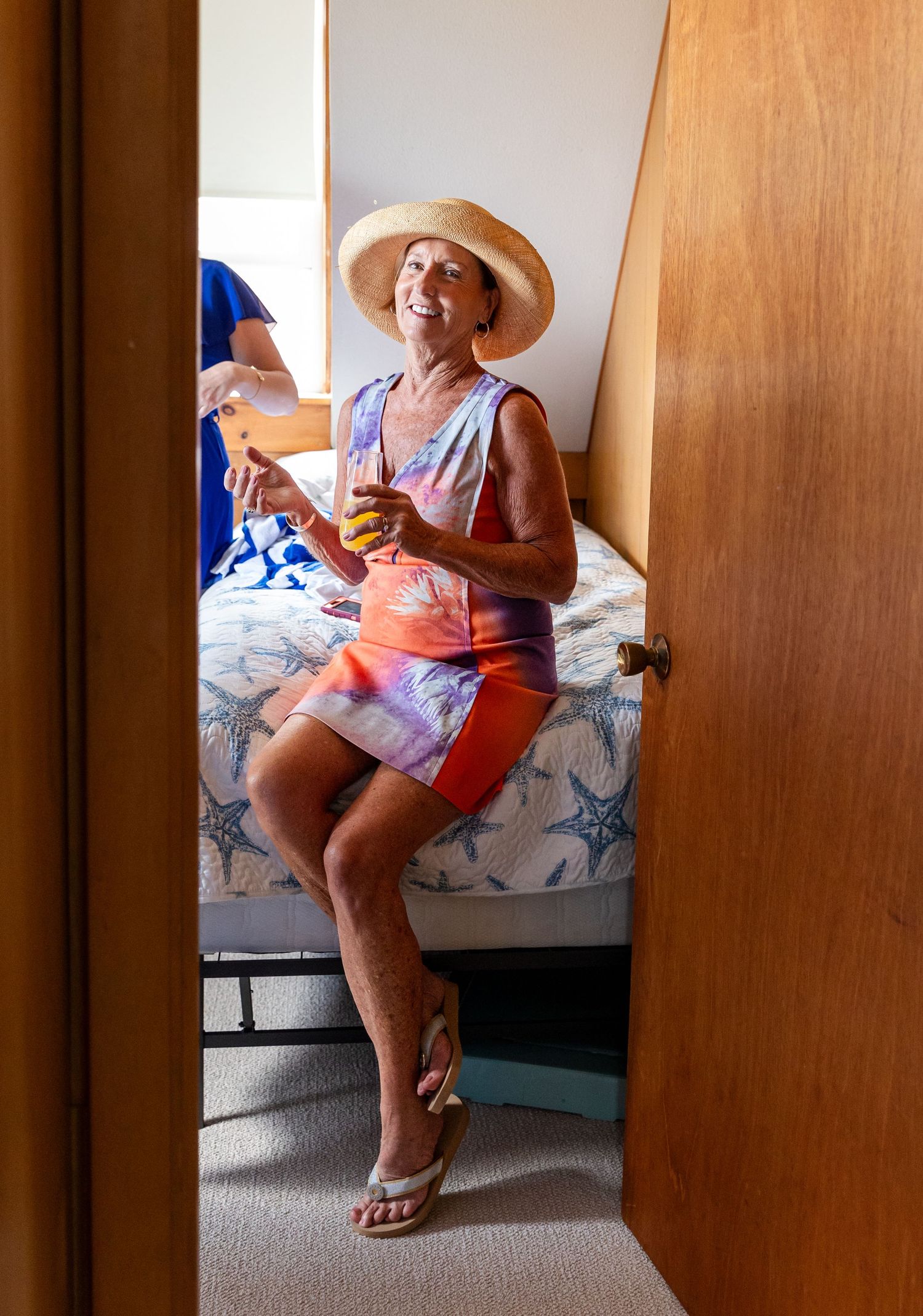 Person in sun hat and colorful summer outfit enjoying a drink while sitting on bed in casual indoor setting.