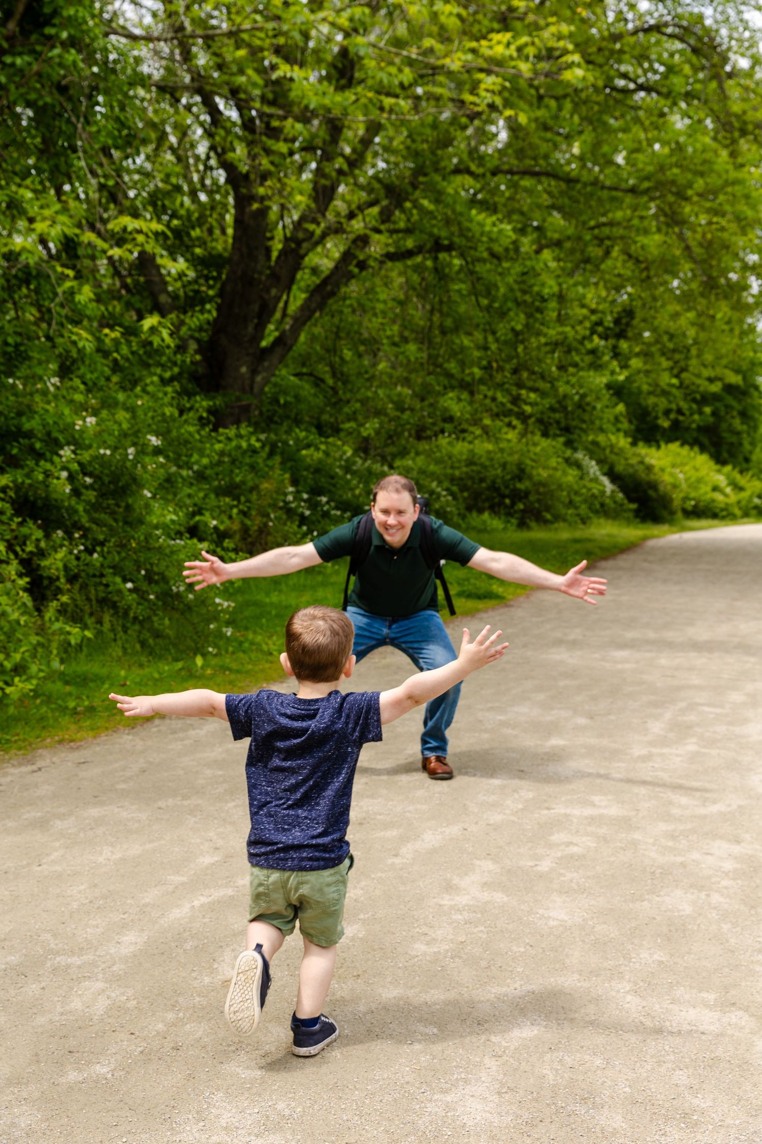 Two people run towards each other on a paved path surrounded by lush green trees in an embrace.