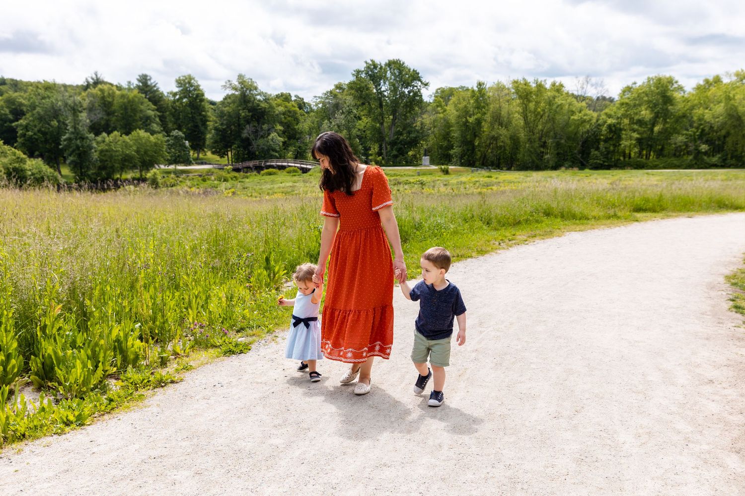 A person in a flowing red dress walks hand in hand with two small children along a gravel path beside a summer meadow.