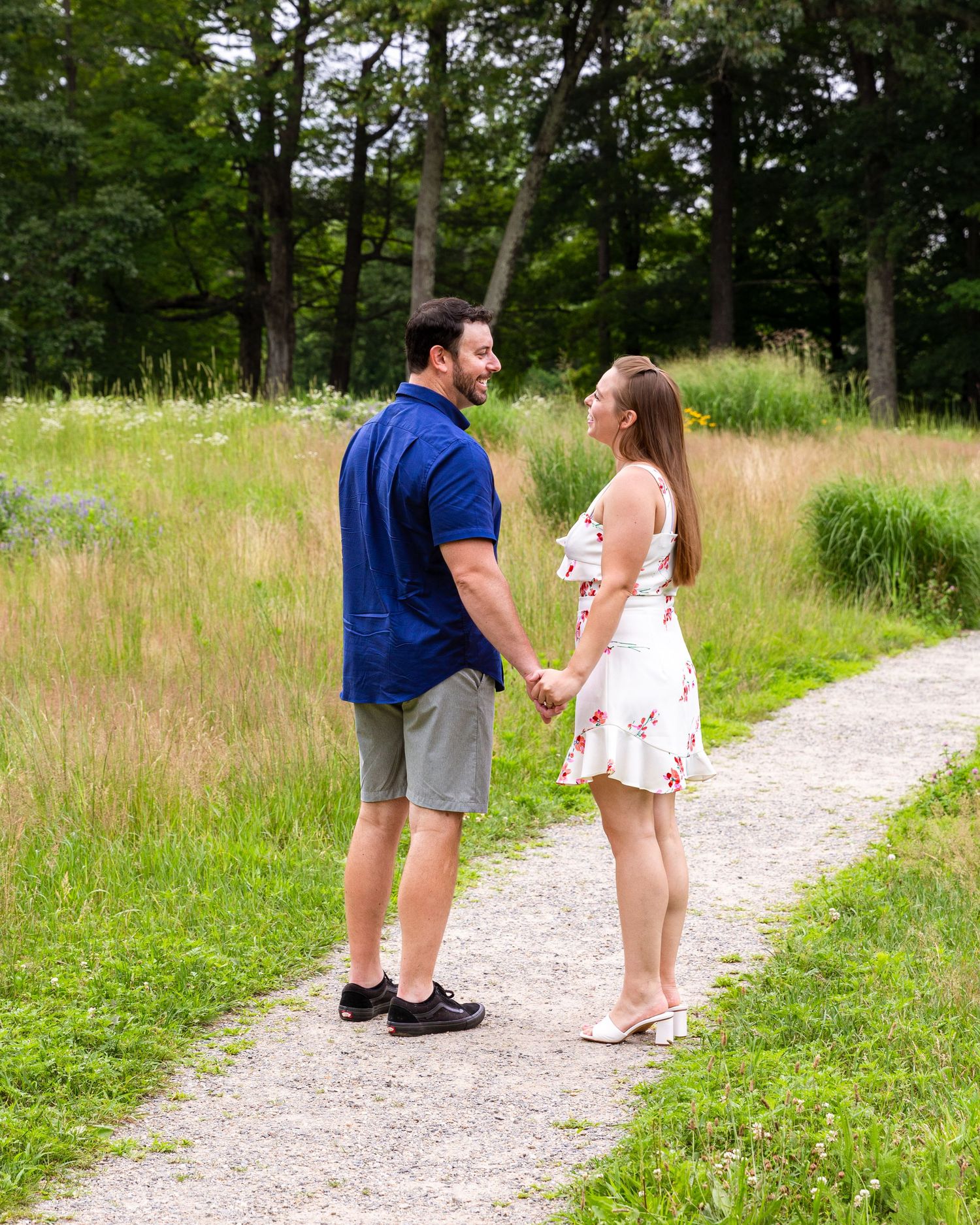 Couple holding hands and smiling while walking on gravel path through tall grass.