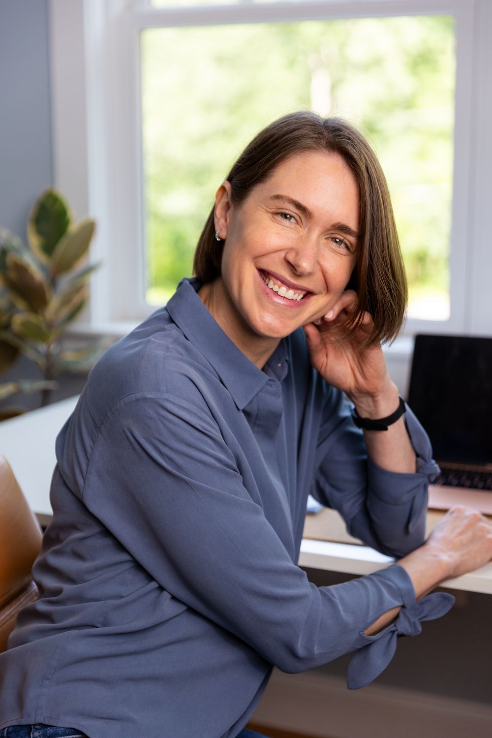 Professional wearing a blue shirt smiles warmly while working at home office desk with laptop and plants.