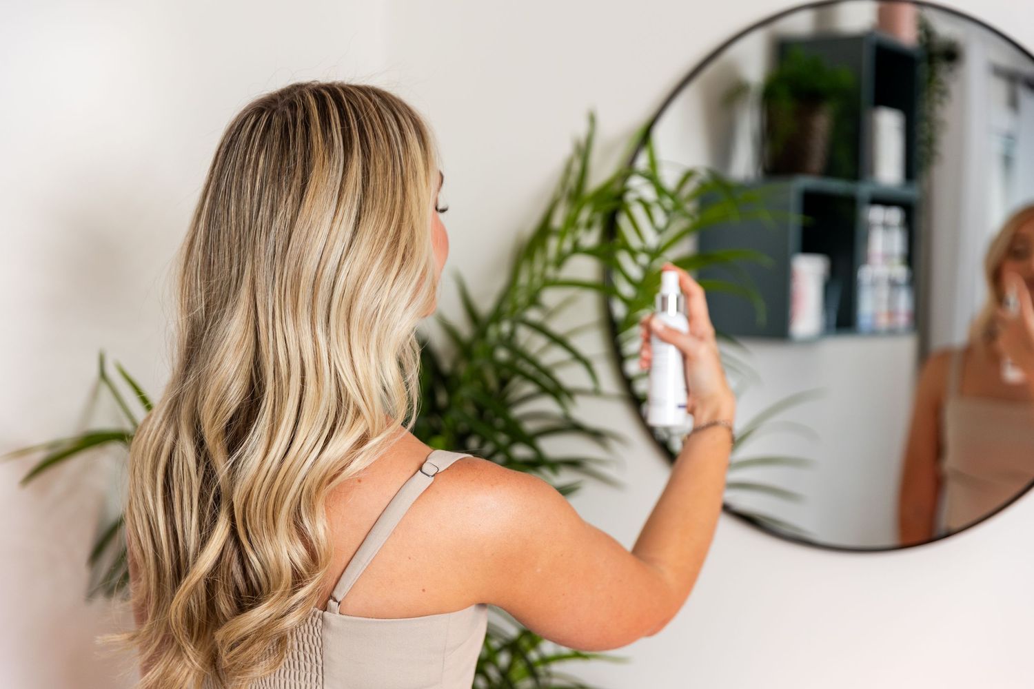 Person with long blonde wavy hair sprays hair product in bathroom mirror with green plants in background.