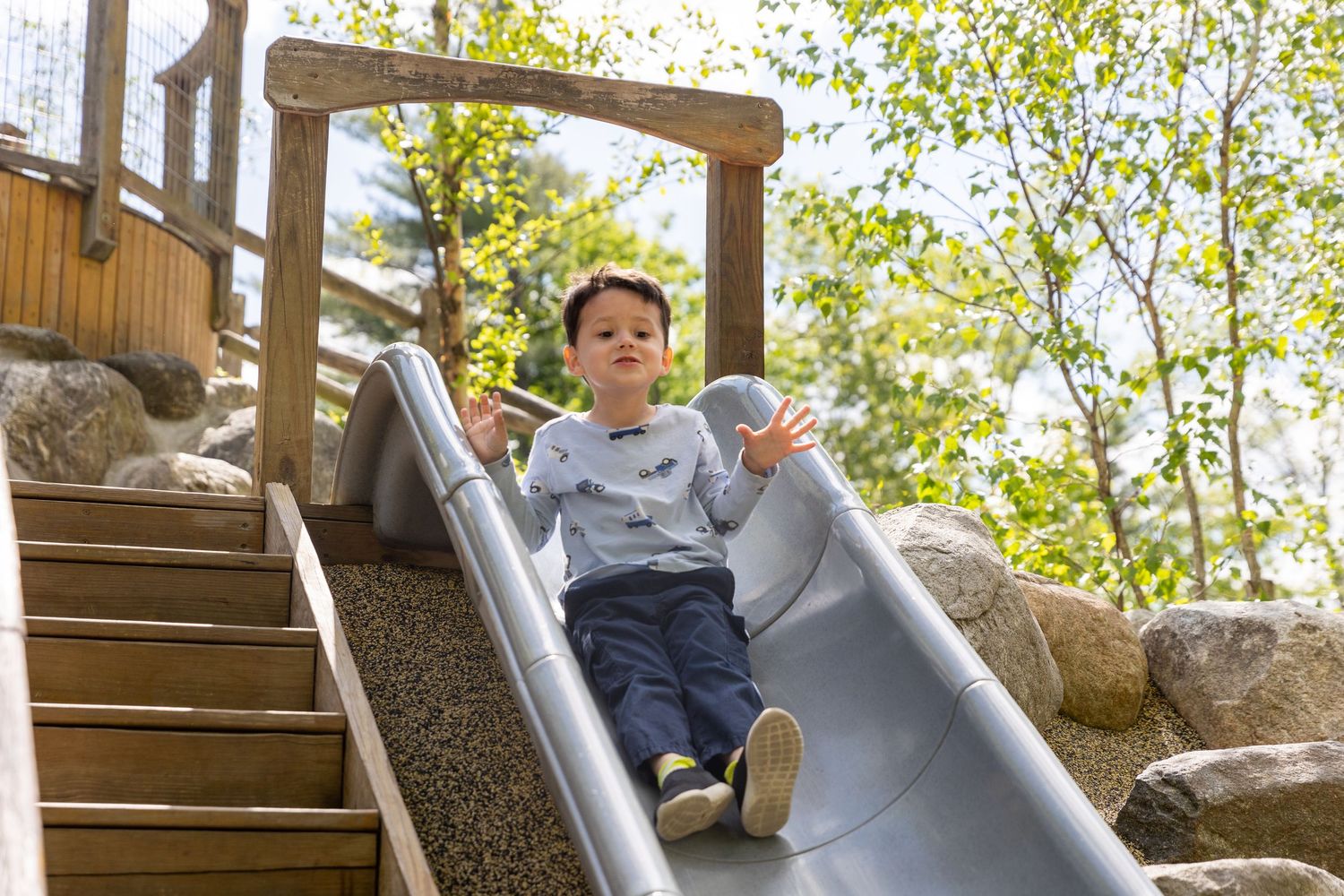 A child slides down a metal playground slide on a sunny day with wooden stairs and rocks visible in the background.