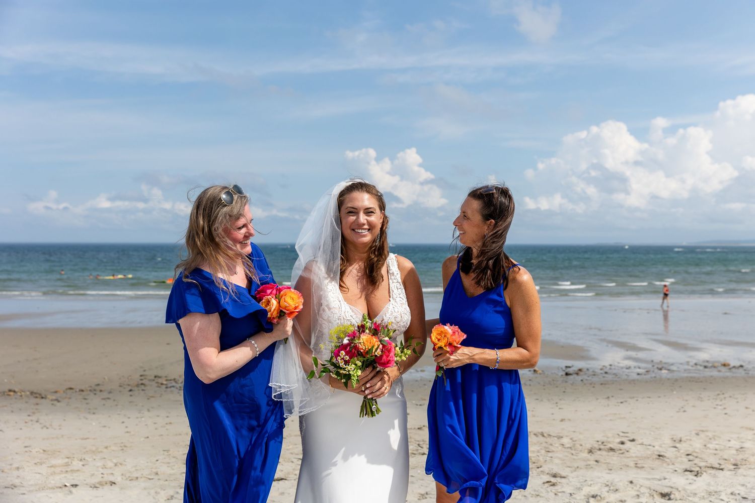Three women in white and navy dresses share drinks and smiles while celebrating at a sunny beach wedding celebration.