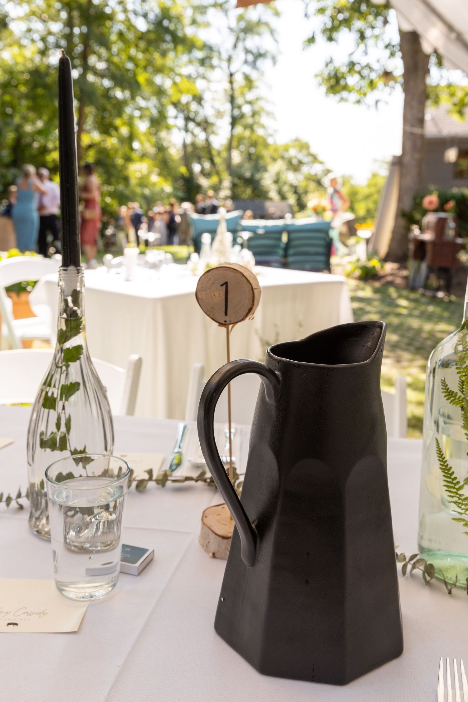 Black ceramic water pitcher sits on white tablecloth at outdoor event with table number and glassware.