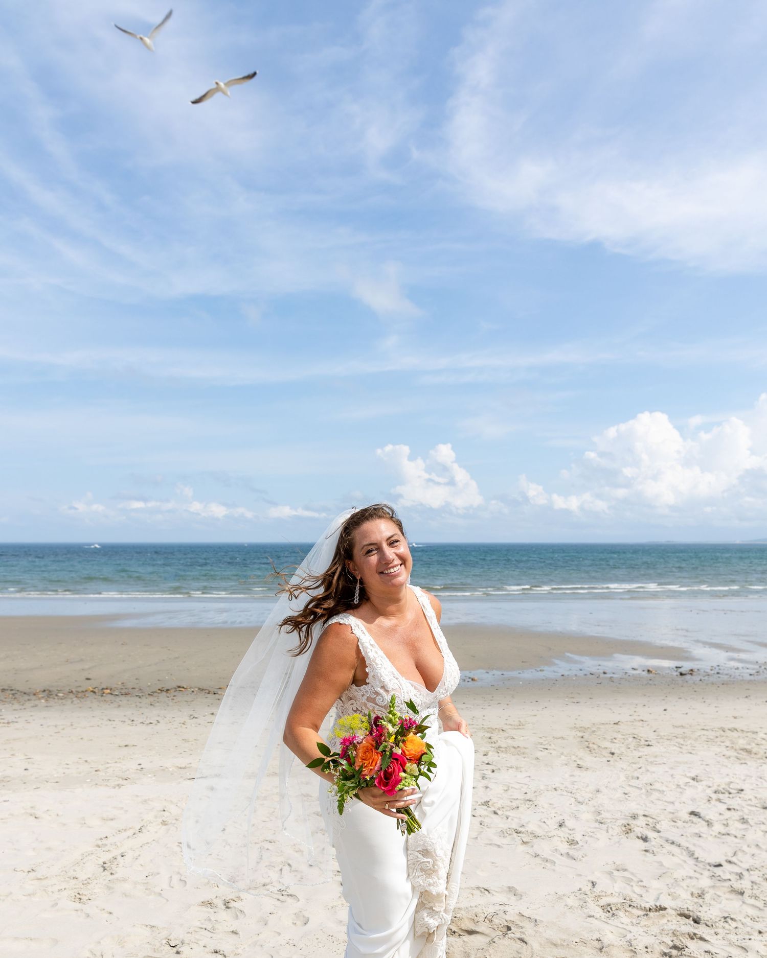 Seabirds soar over a sunny coastal beach scene with white sand, blue waters, and scattered clouds in the sky.