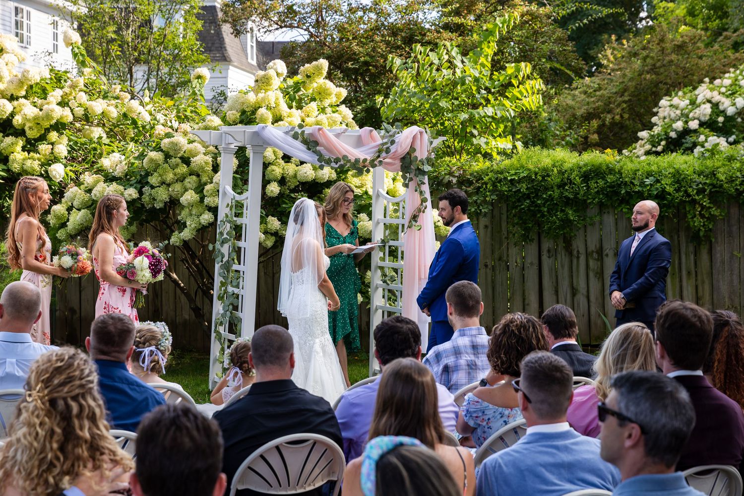 An outdoor wedding ceremony takes place under a white arbor surrounded by blooming hydrangeas and lush summer gardens.
