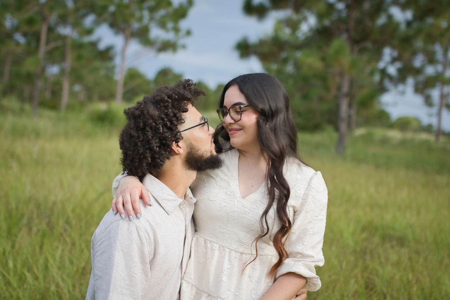 Affectionate couple in matching white outfits smiling and embracing each other in a sunny meadow with trees.