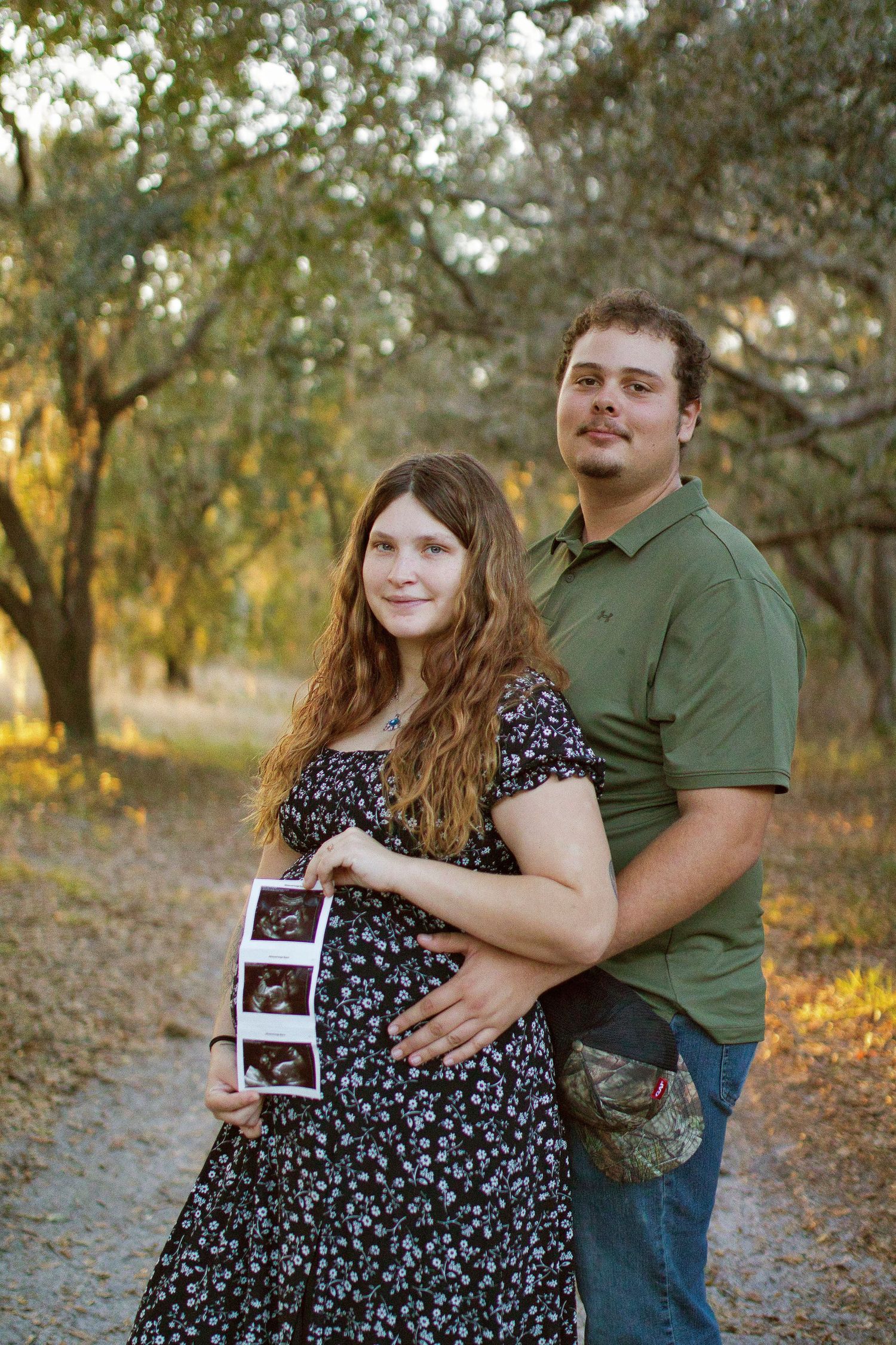 Pregnant couple holding ultrasound photos outdoors on a tree-lined path during golden hour maternity photoshoot.