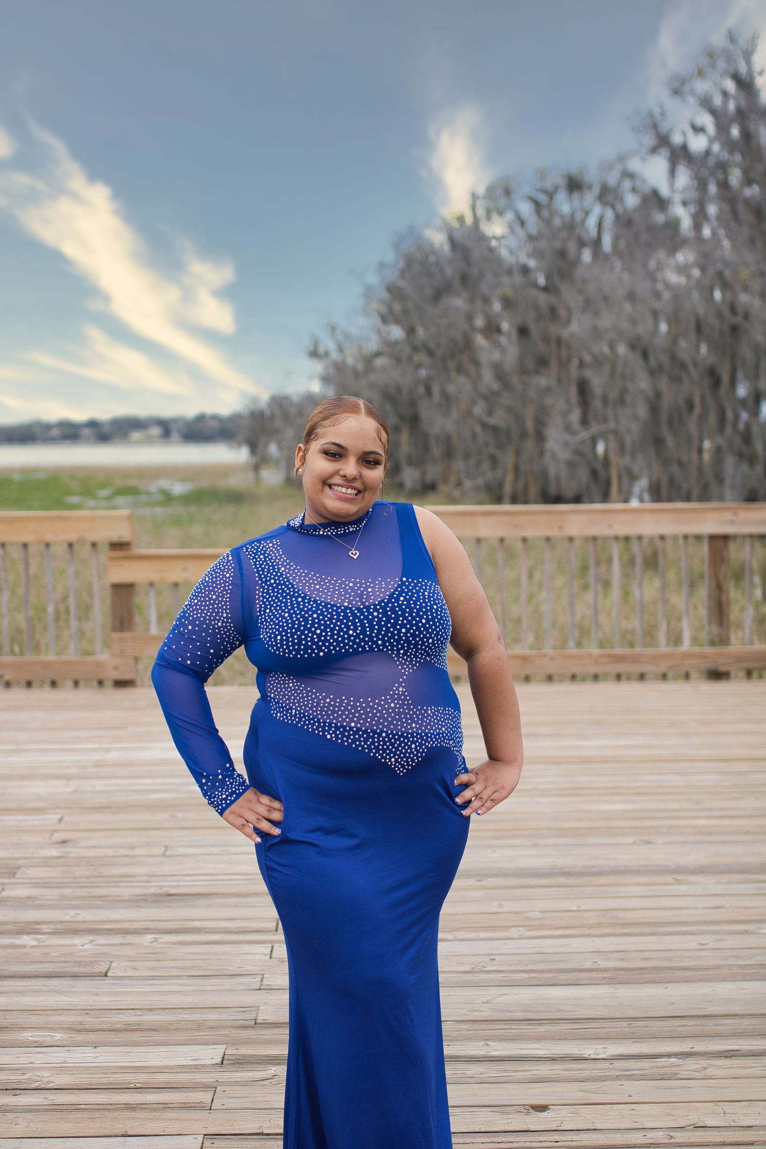 Woman in royal blue rhinestone gown posing on wooden deck near lake with trees and cloudy sky.
