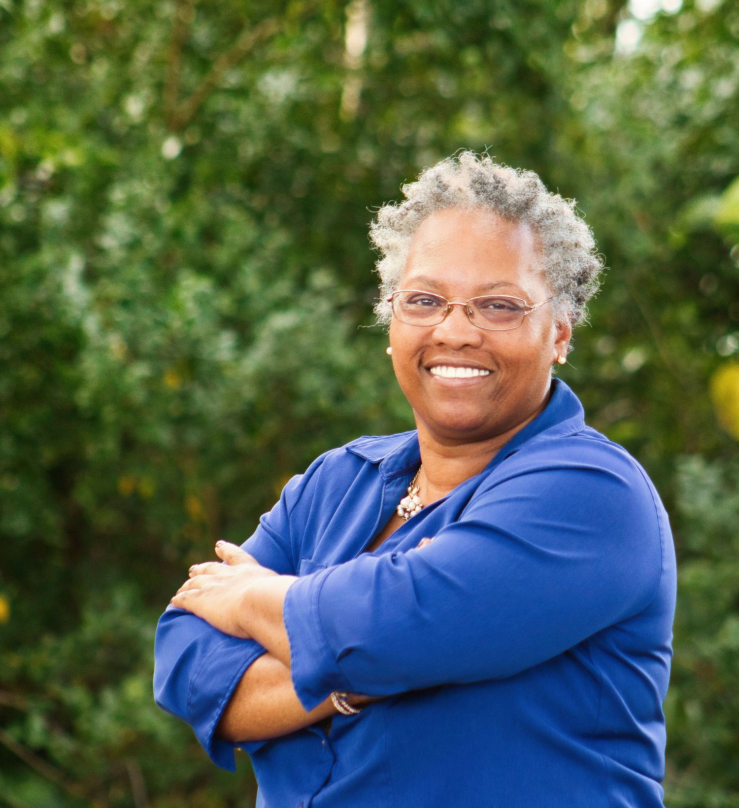 Smiling middle-aged woman with gray hair and glasses, arms crossed, wearing a blue shirt outdoors.