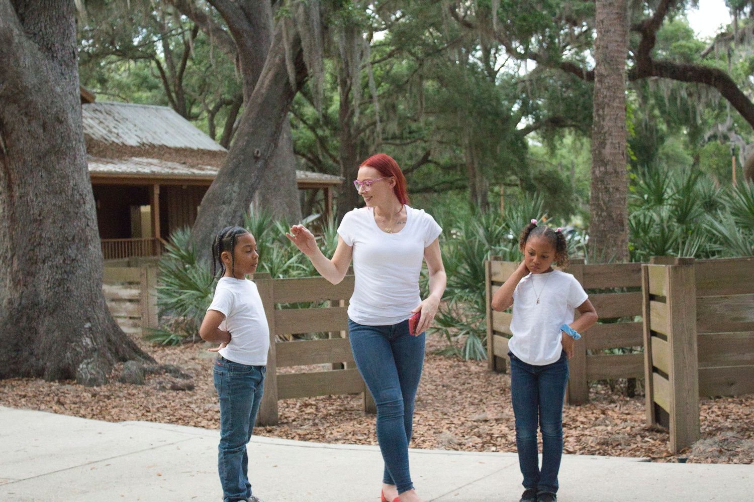Woman with red hair walks with two children outdoors near large oak trees and a wooden fence.