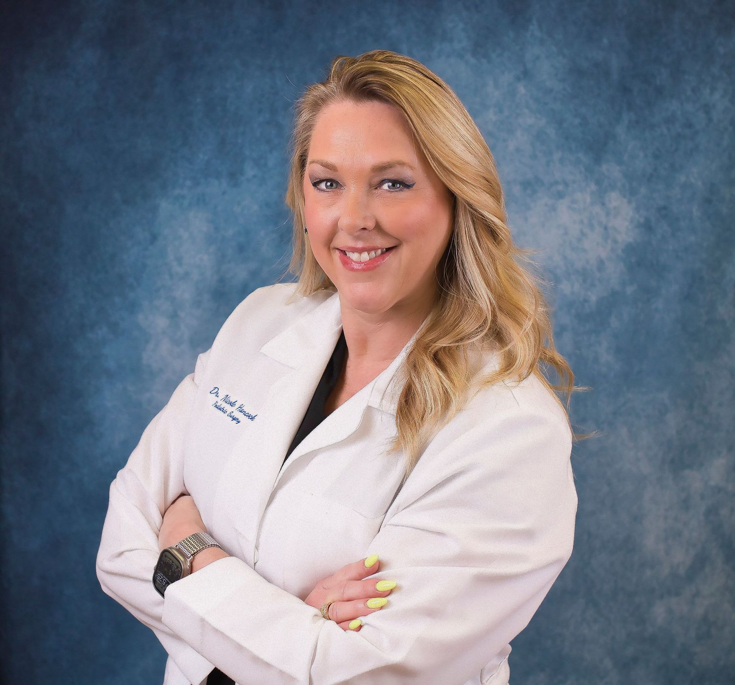 Female doctor in white lab coat smiling confidently with arms crossed against blue background.