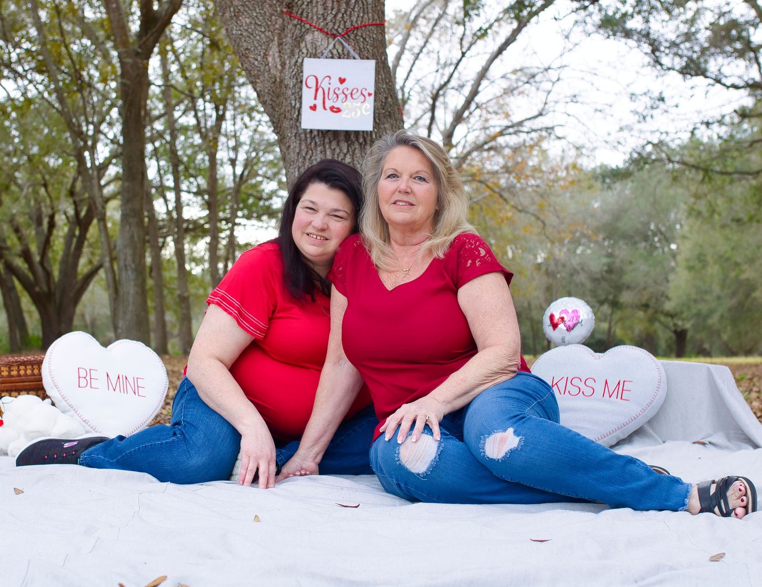 Two people in red tops and jeans sitting on white blanket with Valentine heart pillows in outdoor setting.