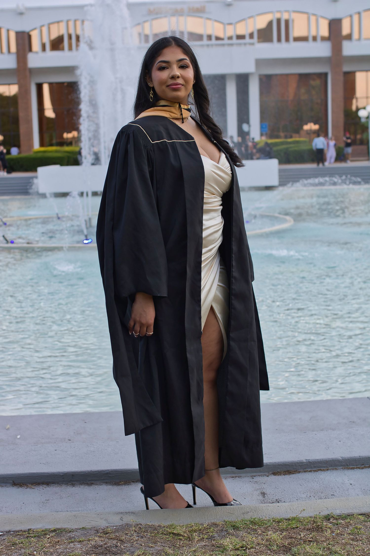 Graduate in black cap and gown poses confidently by fountain wearing cream dress and heels at graduation ceremony.