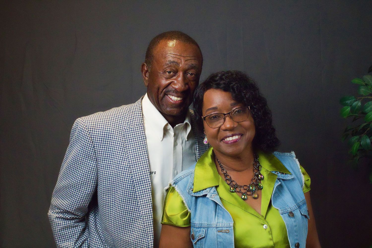 A couple smiles warmly together while wearing business casual attire against a dark backdrop.