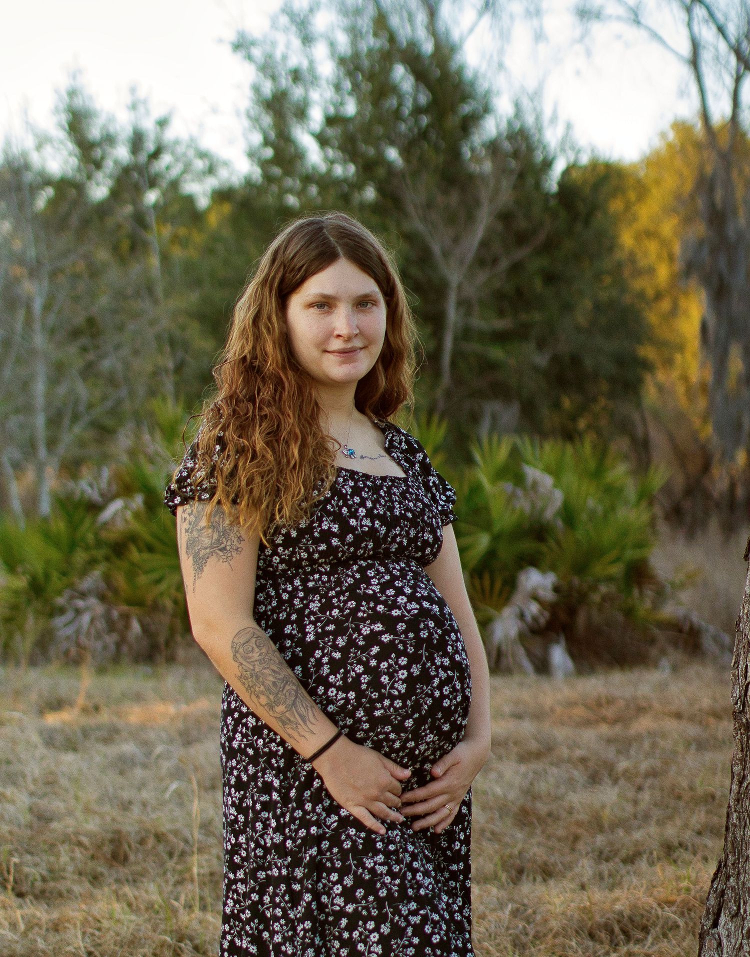 Pregnant woman in floral dress holding baby bump outdoors in natural setting with trees and golden light.