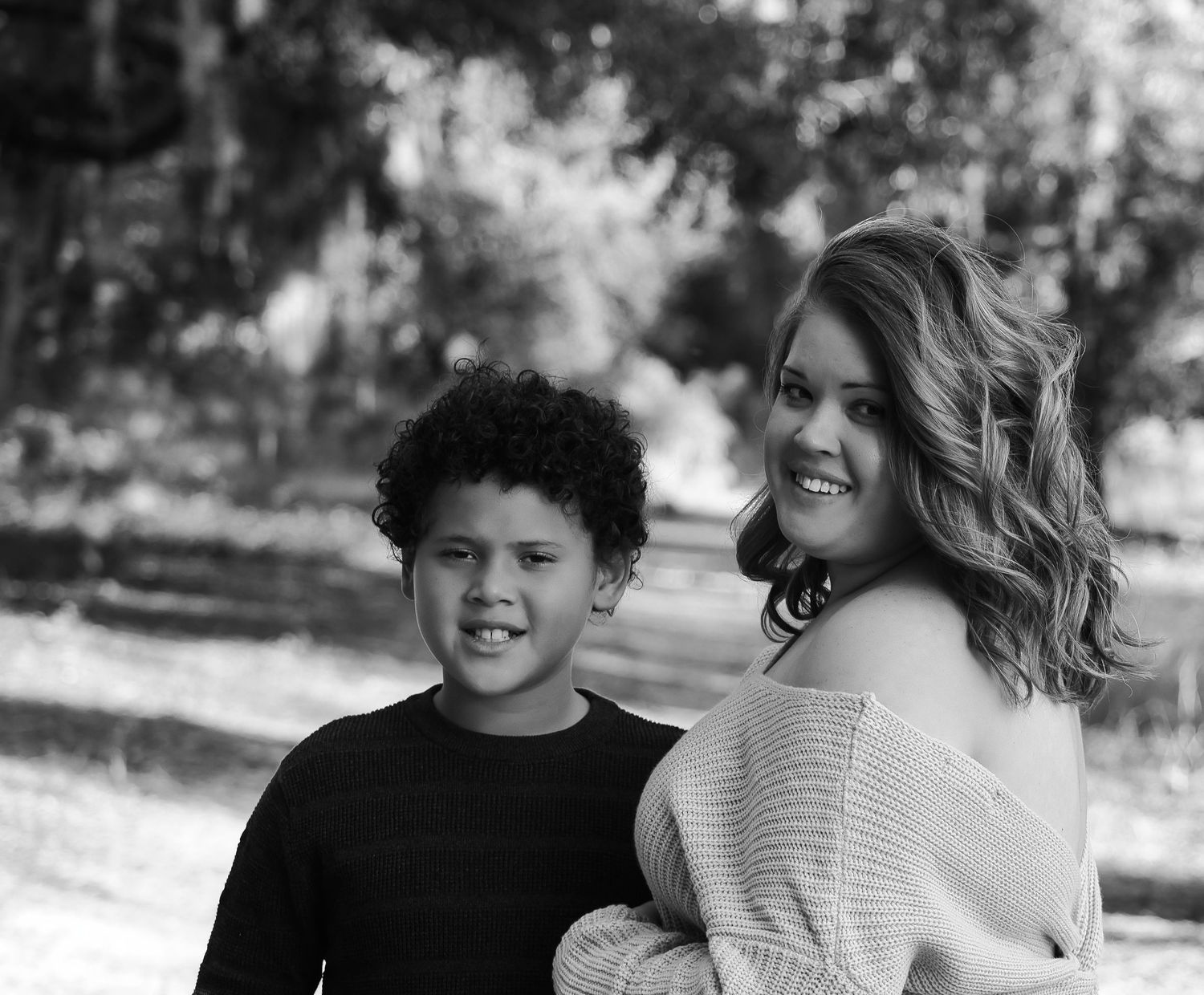 Black and white portrait of a smiling woman and young boy with curly hair posing outdoors near trees.