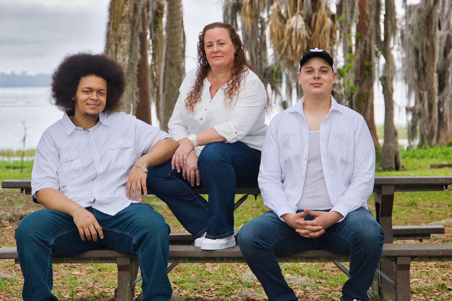 Three people sitting on a bench outdoors near a lake, surrounded by trees, wearing casual white shirts and jeans.