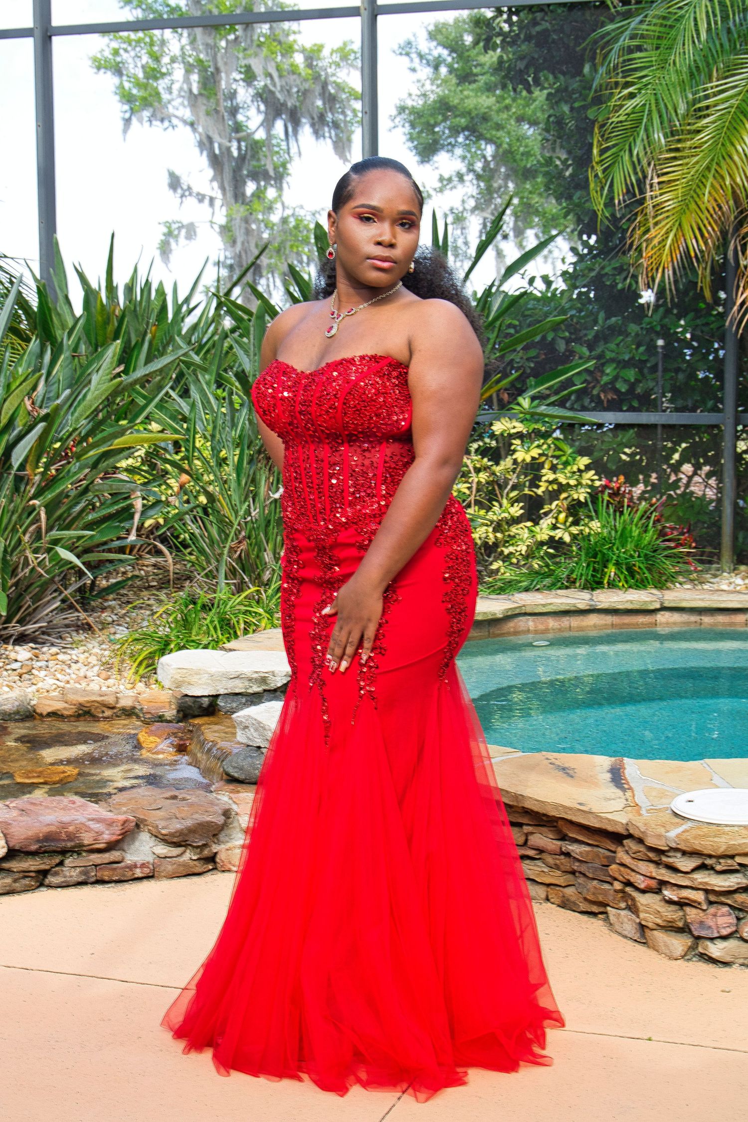 A person in a stunning red sequined mermaid gown poses by a pool surrounded by tropical landscaping.