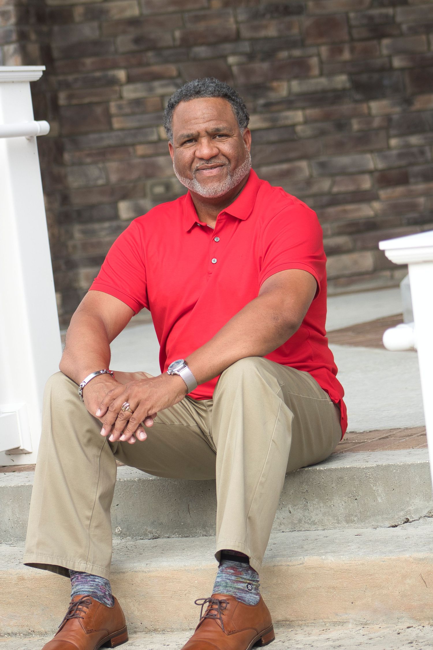 A relaxed individual wearing a red polo shirt and khaki pants sits on steps against a brick wall backdrop.