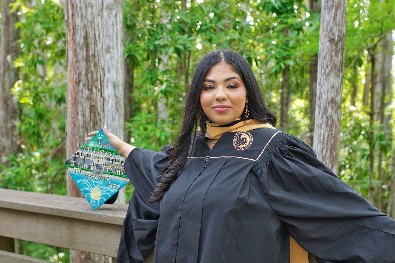 A graduate in academic regalia stands outdoors in a wooded setting holding a decorated graduation cap.