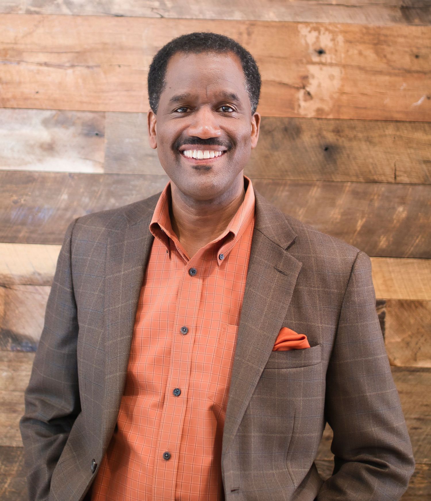 Smiling man in brown plaid blazer and orange shirt with pocket square, posing against rustic wood wall.