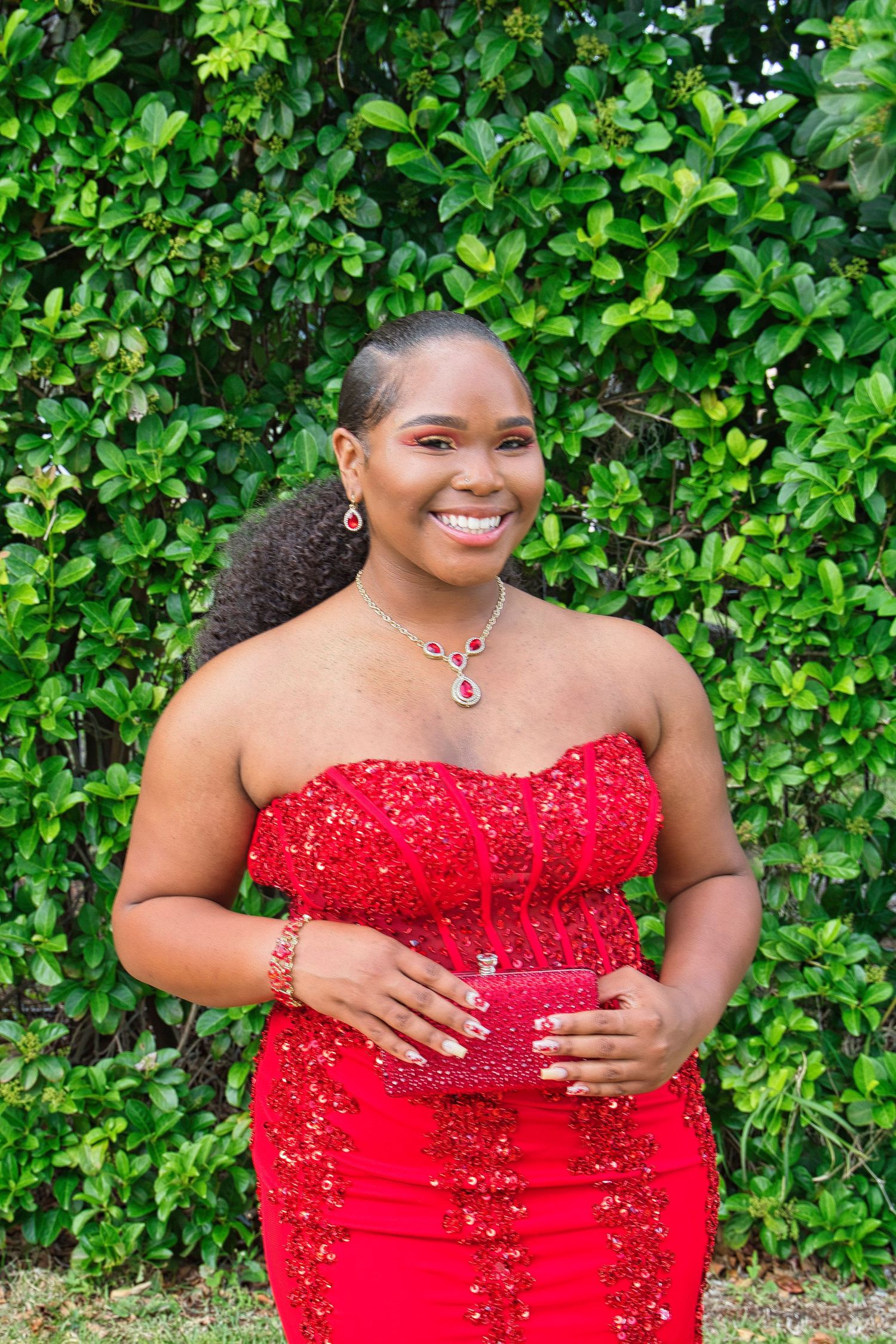 A stunning red beaded mermaid gown is showcased against a lush green garden backdrop.